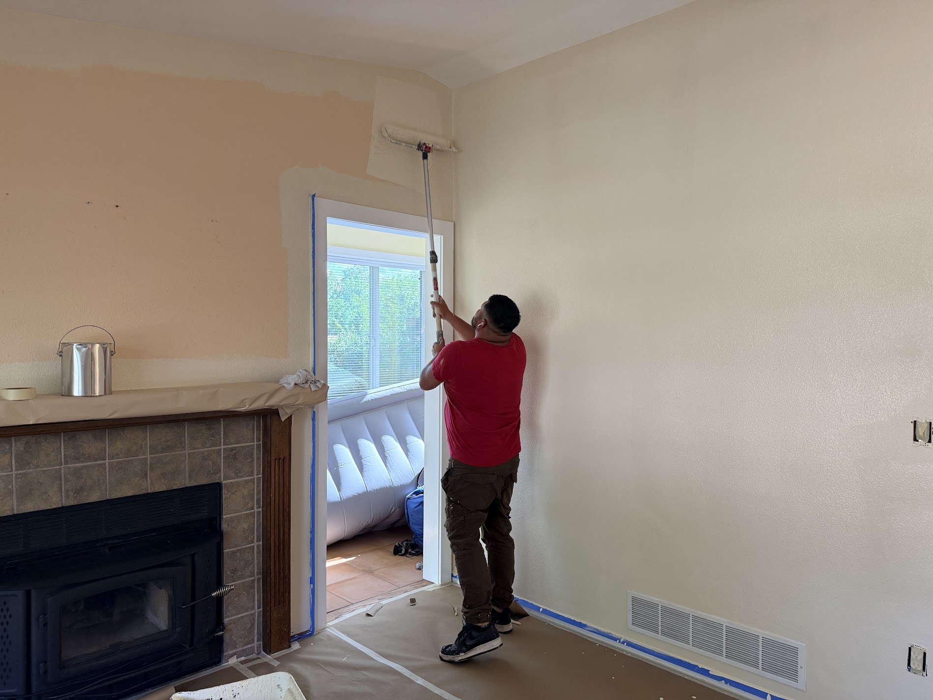 A painter in a red shirt rolling the upper wall of a living room beside a fireplace