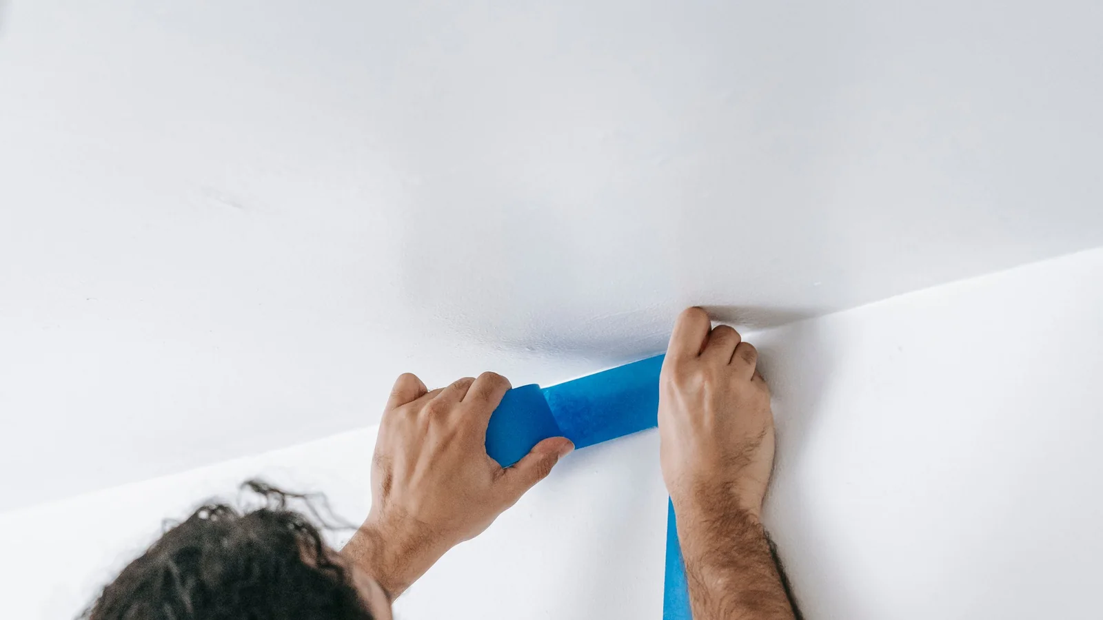 Hands running blue painter's tape along a ceiling line before painting
