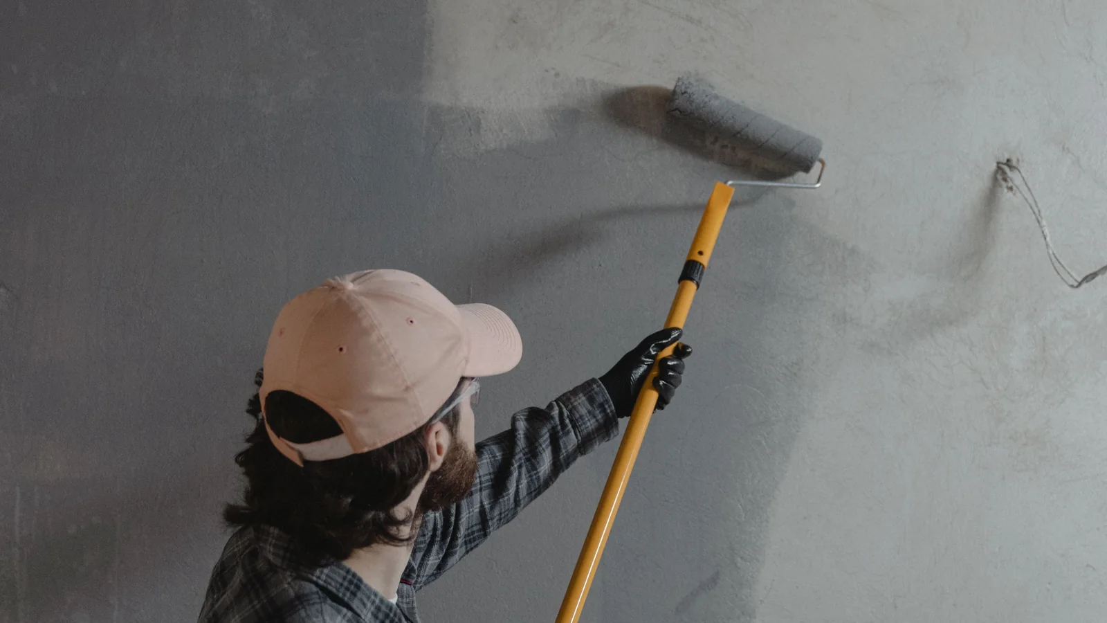 A painter rolling gray paint onto a wall from an extension pole, seen from behind